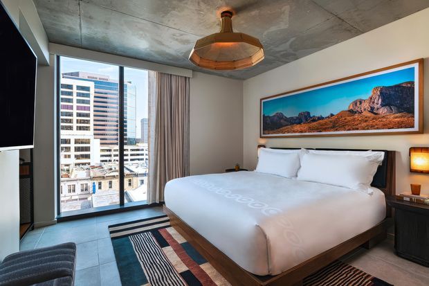 Contemporary urban bedroom with a large platform bed, white bedding, dark timber base, and a colorful framed mountain landscape above the headboard. Floor-to-ceiling glazing offers city views, while concrete ceiling panels, tiled flooring, dark joinery, striped rugs, and a sculptural pendant create an industrial-modern atmosphere.