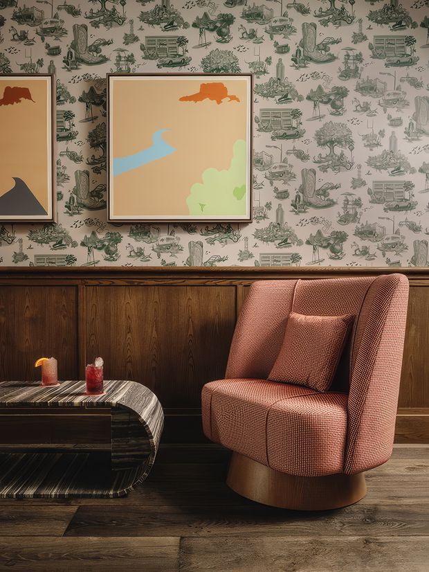 A richly styled living room vignette featuring a sculptural rust-colored upholstered armchair with matching cushion, positioned beside a low rounded dark stone or marble coffee table. The room has dark timber wall panelling and flooring, whimsical illustrated wallpaper, framed abstract artwork, and warm ambient lighting. The overall look is moody, retro-inspired, and refined.