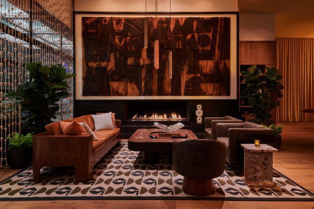 A richly layered, moody living room with a built-in black fireplace wall, linear gas fire, and large framed abstract artwork. The space features a warm timber ceiling and flooring, timber joinery, a patterned area rug, rust-coloured sofa, dark upholstered lounge chairs, a sculptural coffee table, indoor plants, and a decorative beaded screen separating the room from an adjacent area. Warm accent lighting creates an intimate, luxurious atmosphere.