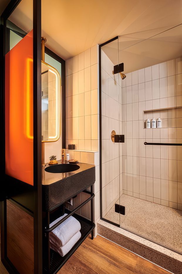 A compact, warmly lit modern bathroom featuring a walk-in glass shower with white square wall tiles, a terrazzo-like speckled shower floor and step, black hardware and grab rail, and a recessed shelf with toiletries. Beside it is a dark textured vanity with a round black basin, copper-toned tap, open black metal shelving, and folded towels. A large illuminated mirror and bold orange translucent panel add a dramatic accent, while the floor has a warm timber finish.