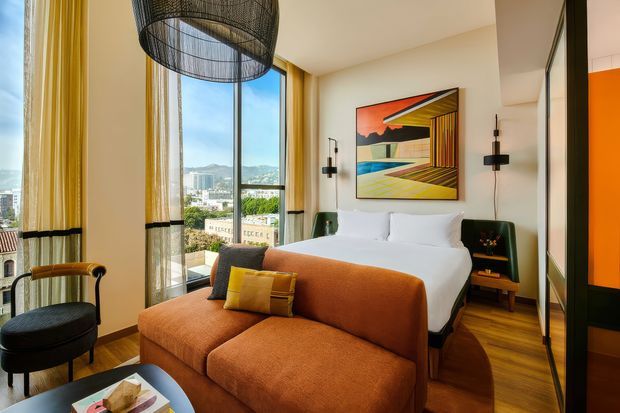 A stylish modern bedroom with a large upholstered rust-orange sofa in the foreground, a neatly made bed, and expansive floor-to-ceiling windows overlooking an urban landscape and hills. Warm wood flooring, cream walls, mustard-yellow curtains, black window framing, sculptural black pendant lighting, and colourful abstract artwork create a vivid, refined interior. A compact black lounge chair and side table sit beside the bed.