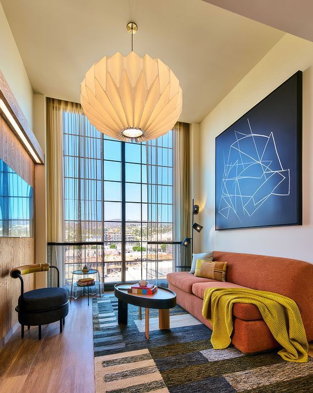 A compact, richly styled living room with a dramatic sculptural pendant light, full-height black-framed windows, warm timber flooring, and a rust-orange sofa draped with a mustard throw. A large geometric artwork hangs above the sofa, while a dark rounded lounge chair and oval coffee table complete the room. The space has abundant daylight, high ceilings, and an urban view beyond the glazing.