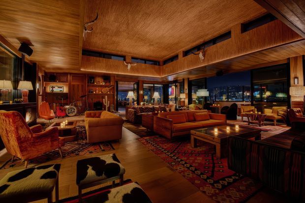 A richly appointed, expansive living room or lounge with a warm wood-lined ceiling and walls, dark timber built-in cabinetry, extensive glazing, and a nighttime city view. Multiple rust, caramel, and brown leather sofas and armchairs are arranged around low timber coffee tables, with layered patterned rugs, decorative lighting, a bar or display area, and a dramatic, intimate atmosphere. The room has a strong mid-century-inspired, lodge-like character with abundant timber and vintage furnishings.