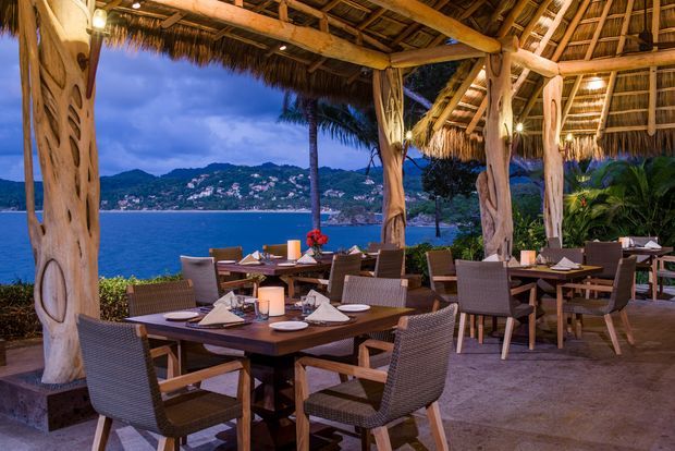 An open-air waterfront dining terrace beneath a rustic thatched roof, supported by substantial natural timber posts and beams. The space contains multiple dark timber dining tables with woven chairs, place settings, candles, and tropical planting. A calm bay, palm trees, and a mountainous coastal settlement are visible beyond the terrace at dusk.