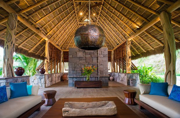 Open-sided tropical pavilion lounge with a dramatic steep thatched timber roof, exposed rafters and heavy rustic posts. A central rough masonry fireplace or feature wall supports a large suspended sculptural metal vessel, while built-in lounge seating, blue cushions, stone surfaces and tropical planting create a relaxed resort-like atmosphere.