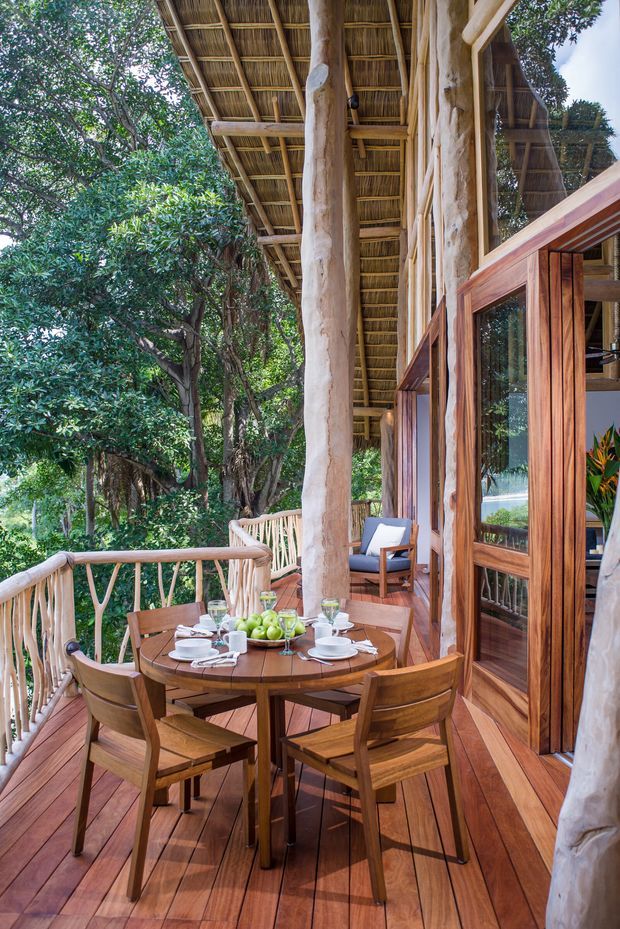 A covered outdoor dining terrace set among dense trees, featuring a round timber dining table with place settings and wooden chairs. The terrace has warm timber decking, organic branch-like posts and balustrades, large timber-framed glazed doors, and a distinctive thatched or woven canopy roof. A lounge chair is visible beyond the dining area.