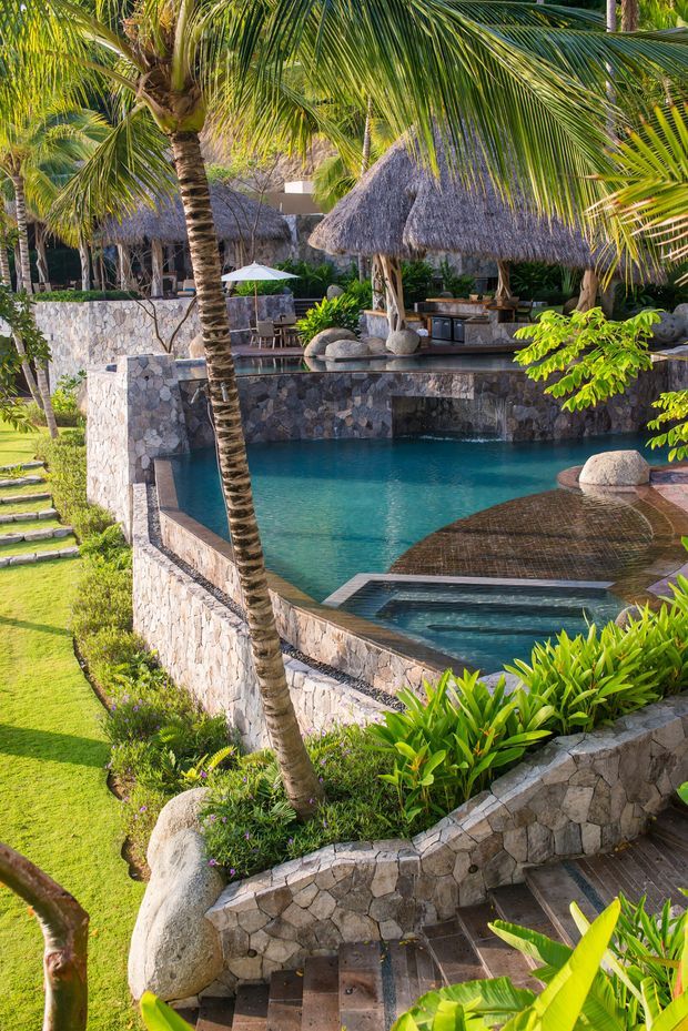 A tropical resort-style outdoor pool area with multiple levels, a turquoise swimming pool, integrated spa or water feature, dark timber decking, and extensive natural stone retaining walls. Dense palms and lush planting surround a thatched-roof poolside pavilion and bar, with stone steps leading through the landscaped garden.