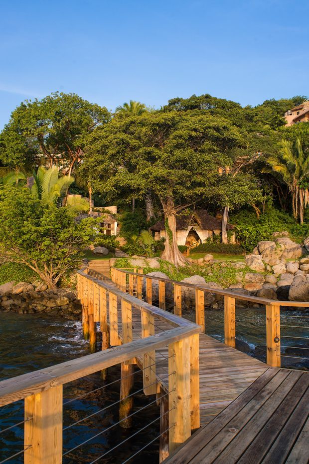 A sunlit timber boardwalk with cable balustrades curves along a rocky waterfront through dense tropical landscaping. A small rustic thatched structure and parts of buildings are partially visible among the trees and rocks.
