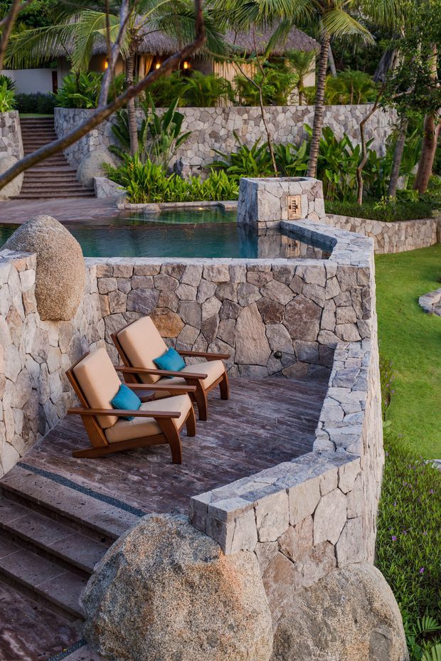 A tropical landscaped outdoor retreat with a small swimming pool, stone retaining walls, stepped terraces, lush palms and planting, and two wooden lounge chairs with teal cushions. The space has a resort-like atmosphere and extensive natural stone masonry integrated into the garden and pool area.