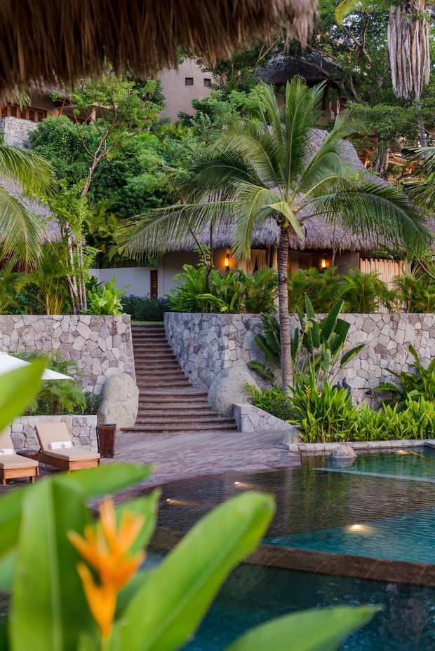 Lush tropical resort-style outdoor setting with a stepped swimming pool, stone retaining walls and stairs, dense palms and tropical planting, and thatched-roof structures surrounded by greenery. Warm lighting and natural stone create an exotic, tranquil atmosphere.