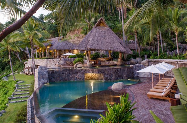 Tropical resort-style outdoor pool area surrounded by lush palms and dense greenery. The scene features a stone-edged swimming pool with integrated lighting, a thatched-roof pool bar or pavilion, timber decking, lounge chairs, white umbrellas, stepping-stone paths, and warm evening lighting.