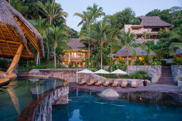 Tropical resort-style property with multiple thatched-roof structures, lush palm landscaping, stone retaining walls, and a large curving swimming pool surrounded by sun loungers and umbrellas. The image is an exterior architectural and pool-area view photographed at dusk.