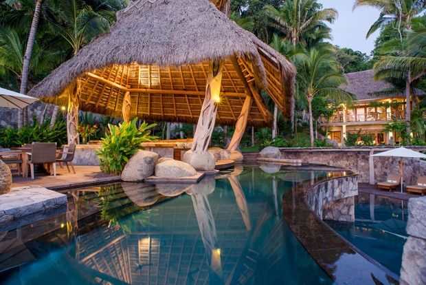 A tropical resort-style outdoor pool area dominated by an open-sided thatched pavilion with a dramatic pitched roof, natural timber supports, warm lighting, lush palms, stone landscaping, and poolside seating. A multi-level resort building is visible in the background.