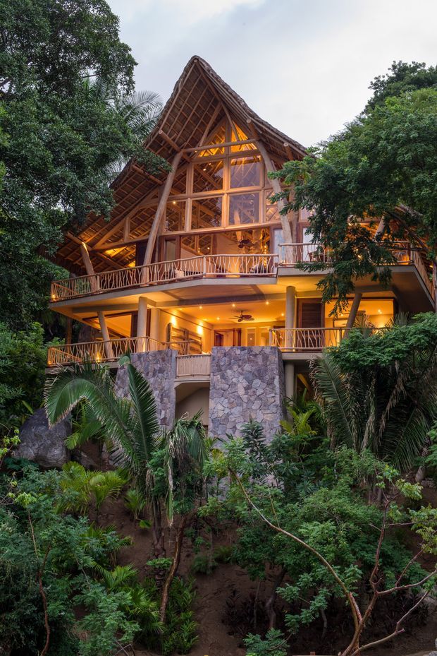 A multi-level tropical house nestled in dense lush vegetation. The building features dramatic steeply pitched gabled roofs with exposed timber rafters, extensive timber-framed glazing, open balconies with rustic branch-style balustrades, warm evening lighting, and substantial natural stone-clad lower walls and columns.