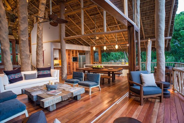 Open-plan tropical living and dining space on a timber deck, featuring a high exposed timber structure, natural tree-trunk posts, a woven thatched ceiling, extensive open glazing, timber flooring, relaxed upholstered seating, a rustic coffee table, and a dining table adjacent to a compact kitchen. Lush greenery is visible beyond the open sides.
