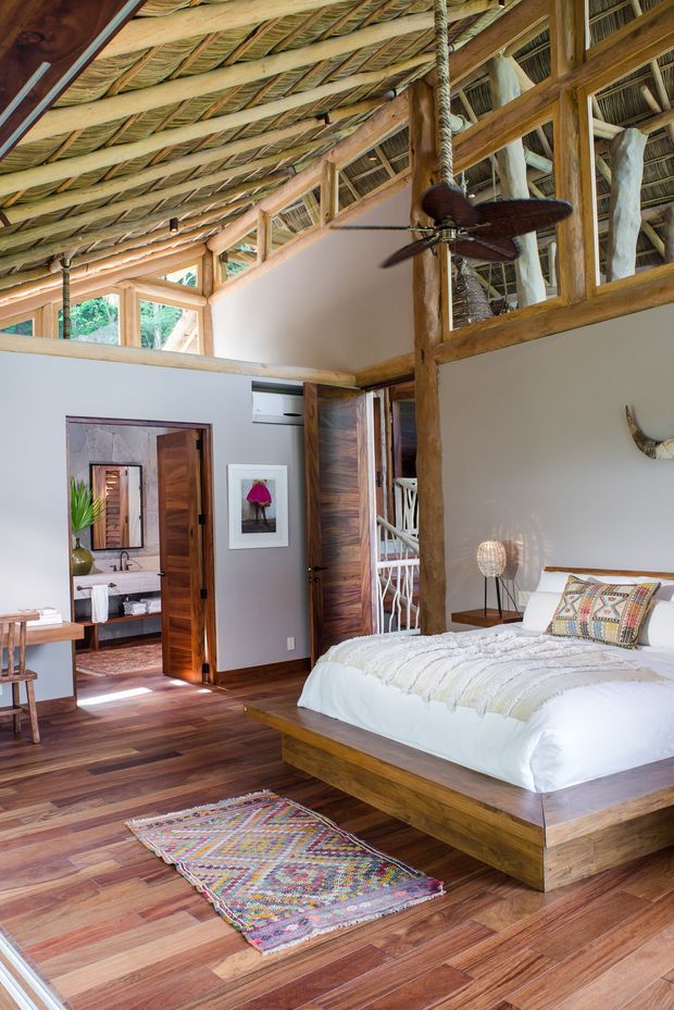 A warm, rustic bedroom with a timber platform bed, white bedding, colourful woven rug and patterned cushions. The room features extensive exposed timber framing, a high pitched thatched or woven ceiling, a ceiling fan, timber flooring, and an open mezzanine-like upper wall with woven infill. A bathroom is visible through the open wooden door, with an additional outdoor or circulation area beyond another opening.