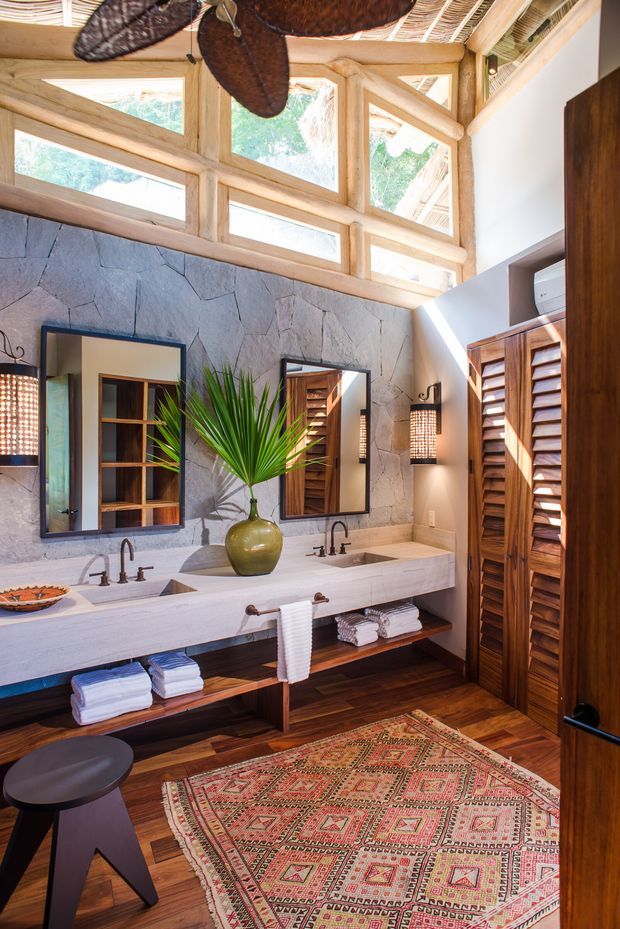 A richly detailed bathroom with a long double vanity, two vessel-style sinks, dark metal tapware, open timber shelving, and folded towels. The walls feature large irregular grey stone slabs, while warm timber flooring, louvred timber cabinetry, exposed timber framing, and high clerestory windows create a bright, natural setting. Woven pendant lights, mirrors, a patterned rug, and tropical greenery add an earthy resort-like character.