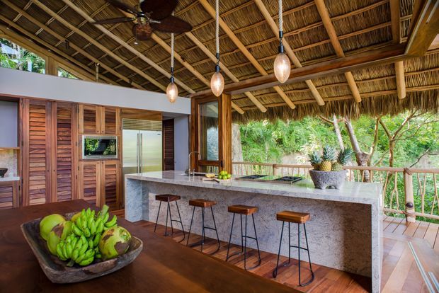 Open-air tropical kitchen and dining area beneath a richly textured thatched roof. The space features a large stone island with timber-topped stools, timber cabinetry and wall panelling, hanging pendant lights, ceiling fans, and a timber dining table. A covered deck with natural timber railings and dense greenery beyond creates a strong indoor-outdoor connection.