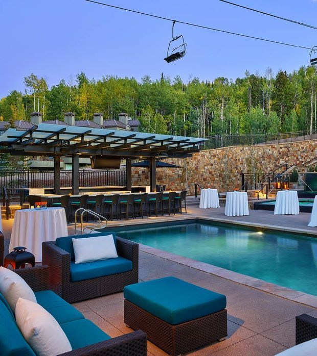 An upscale outdoor pool terrace in a wooded mountain setting, featuring a turquoise swimming pool, dark woven lounge seating with teal cushions, cocktail tables dressed in white linens, and a covered outdoor bar with a translucent roof canopy. A stone retaining wall, railings, resort-style lighting, and overhead chairlift cables are visible in the background.