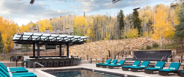 Outdoor pool area with turquoise lounge chairs, a dark tiled swimming pool, and a covered poolside bar or entertaining pavilion. A substantial stone retaining wall and black metal railings frame the space, with dense autumn woodland beyond.
