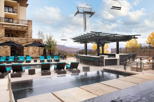 Outdoor resort-style pool terrace beside a multi-storey stone-clad building, with turquoise sun loungers, umbrellas, a long outdoor bar beneath a dark pergola, and a chairlift infrastructure overhead. Mountainous scenery and autumn trees are visible beyond the balustrade.