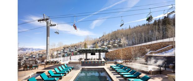Outdoor resort pool area beneath a chairlift, with a rectangular pool, rows of turquoise loungers, a raised bar or service pavilion, and a wooded mountain slope in the background. Steam rising near the pool suggests a heated spa or thermal resort setting. Bright alpine conditions and strong turquoise accents create a lively, recreational atmosphere.