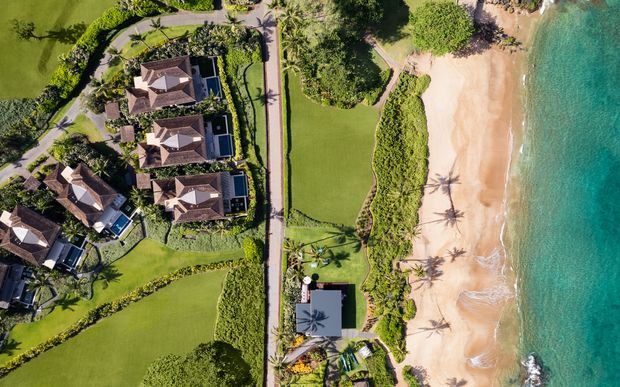 Aerial view of a landscaped beachfront property, showing multiple tiled-roof villas, manicured lawns and gardens, a central access road, and a broad sandy beach beside turquoise water.