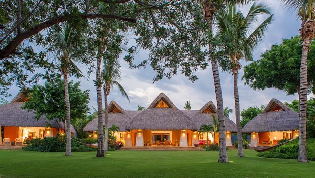 A tropical resort-style property viewed from a manicured lawn, with several low-rise buildings featuring steep thatched roofs, triangular dormer-like gables, warm illuminated interiors, and abundant palm trees and lush landscaping.