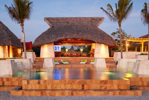A tropical resort-style pavilion or villa with a broad layered thatched roof, open glazed living area, warm interior lighting, palm trees, and a large reflective pool with stone-clad steps and terraces in the foreground. The architecture has strong Polynesian or tropical influences and is presented at dusk.