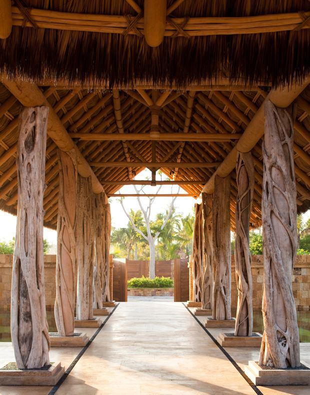 A covered tropical architectural passage or entry pavilion framed by heavily textured natural timber tree-trunk columns. The structure features an elaborate exposed timber post-and-beam roof, layered bamboo or timber rafters, and a thick thatched roof with deep overhangs. A pale stone or tile walkway leads through the pavilion toward a landscaped courtyard and open garden beyond. The composition is symmetrical, warm-toned, and strongly focused on the rustic structural detailing.