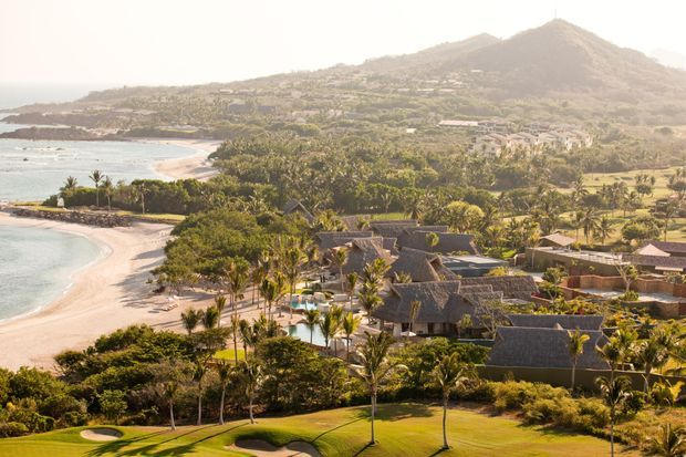 A tropical beachfront resort or residential compound viewed from an elevated position, with curved sandy beaches, turquoise water, dense palm landscaping, multiple low-rise buildings with steep thatched roofs, and a mountainous island backdrop.