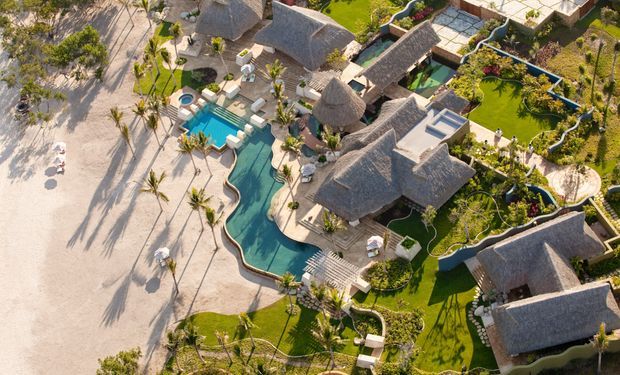 Aerial view of a tropical beachfront resort or residential compound with multiple thatched-roof buildings, curving turquoise swimming pools, manicured gardens, palm trees, paved paths, and sandy beach frontage.