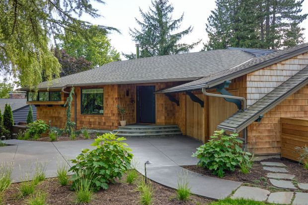 Exterior view of a low, single-storey house with warm cedar shingle cladding, a broad grey asphalt-shingle hipped roof, dark-framed windows and door, and a covered entry porch. Curved concrete paths and landscaped planting lead to the entrance, with mature trees surrounding the property. A smaller attached or adjacent wing with a sloping roof is visible on the right.