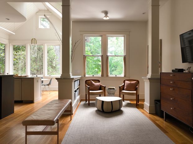 Bright open-plan interior combining a living room and kitchen. The living area features warm timber flooring, a large neutral rug, two leather lounge chairs, a round sculptural coffee table, a leather bench, built-in columns, and a dark timber chest of drawers with wall-mounted television. The adjacent kitchen has light cabinetry, pale benchtops, extensive glazing, and a high partially vaulted ceiling. Large windows overlook leafy greenery, creating an airy, warm, refined atmosphere.