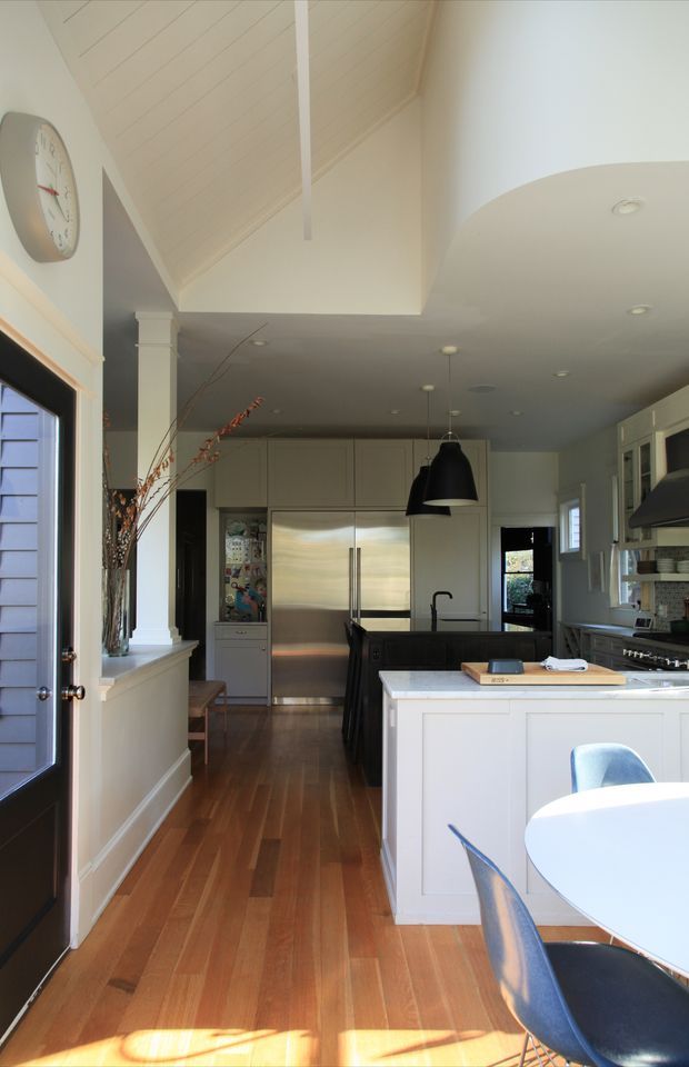 A bright, traditional-style kitchen viewed from the dining area, featuring a high vaulted white ceiling, white built-in cabinetry, stainless-steel double-door refrigerator, dark central island, white island benchtop, pendant lighting, timber flooring, and a partially visible round dining table with blue chairs. Natural light enters through the glazed door and windows.