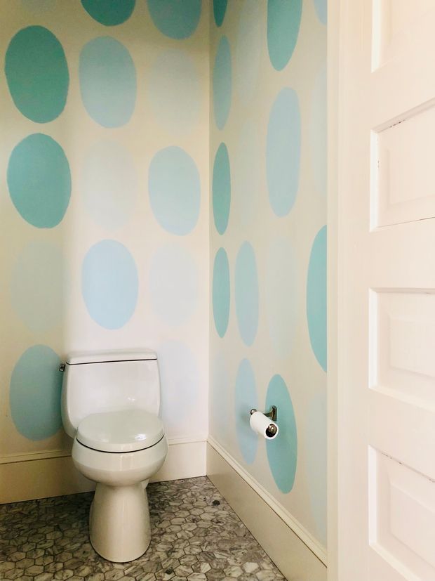 A compact powder room with a white toilet, white trim and paneled door. The walls feature a playful oversized oval pattern in pale aqua, teal and cream, while the floor is covered in small irregular grey-green stone-look tiles. The room has a bright, whimsical, child-friendly character.