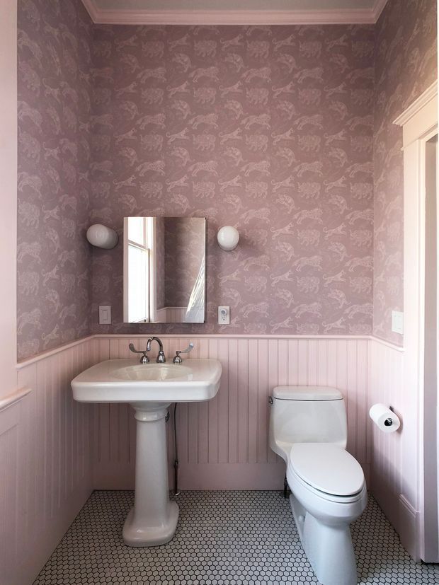 A compact powder room with a pedestal sink, toilet, rectangular mirror, and globe wall sconces. The walls feature pink patterned wallpaper above pink vertical beadboard wainscoting, with white traditional trim and a small hexagonal mosaic tile floor.