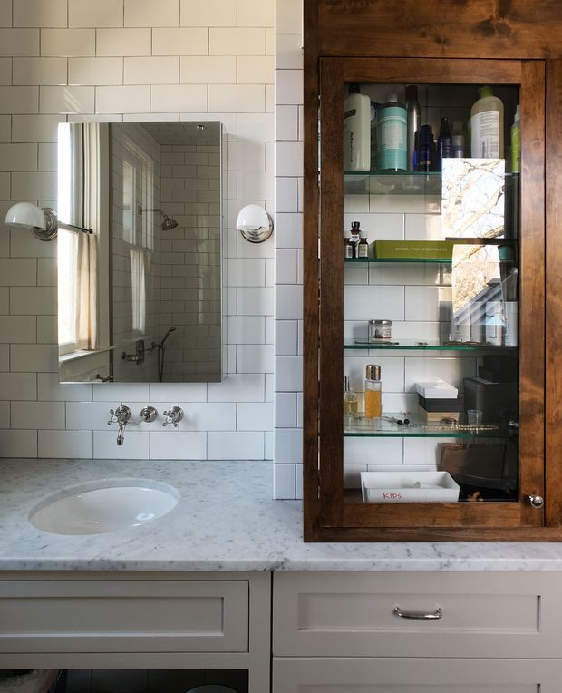 A close-up view of a traditional bathroom vanity. White rectangular wall tiles surround a marble-look countertop with an integrated white basin, wall-mounted chrome tapware, and a rectangular mirror. A dark timber-framed glass-fronted medicine cabinet provides storage beside the mirror, with wall sconces on either side of the mirror. The cabinetry below is painted off-white with recessed panel fronts and chrome handles.