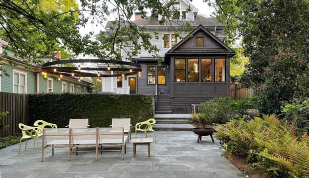 A landscaped residential courtyard and paved patio viewed from outdoors, featuring a dark grey timber-clad enclosed porch or extension with glazed windows, steps, and a pitched gable roof. The patio has a dining table and chairs, pale green accent chairs, a pergola with string lights, clipped hedging, mature planting, and a larger white weatherboard house visible behind.