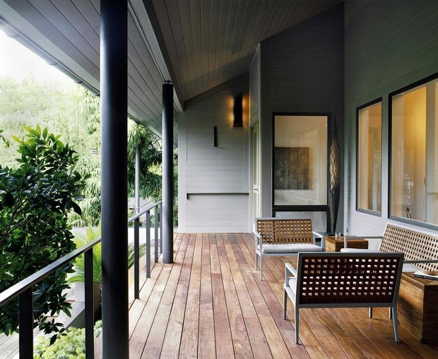 A covered outdoor verandah or balcony terrace with timber decking, dark cylindrical support columns, pale horizontal wall cladding, and a slatted ceiling. Woven lattice outdoor seating and framed glazing line the house, with lush vegetation visible beyond the railing. The space has a refined, warm contemporary character.