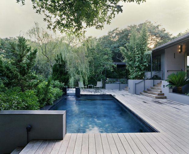 A landscaped residential outdoor area featuring a long rectangular swimming pool surrounded by a broad timber deck. Steps lead up to a contemporary house with a dark roof, pale grey exterior walls, covered porch, and potted planting. Dense mature greenery creates a secluded garden setting.