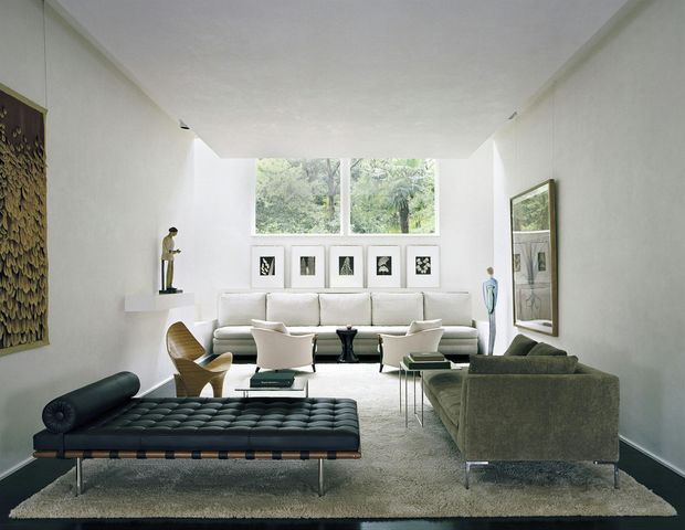 Bright, elongated living room with a high flat ceiling and large rear window overlooking greenery. The room features white walls, a pale built-in or freestanding sofa, sculptural lounge chairs, a dark tufted leather bench, a dark green sofa, framed artwork, and minimalist display objects. The composition has clean modernist lines, balanced natural light, and a refined mid-century-inspired furniture arrangement.