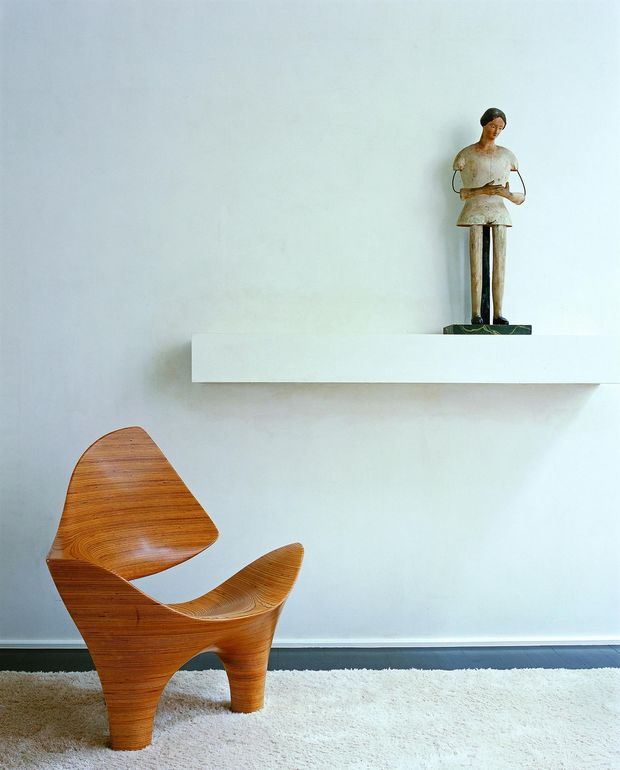 Minimalist interior vignette with a sculptural bentwood lounge chair on a pale textured rug, set against a white wall. A simple floating white shelf displays a small figurative sculpture, creating a sparse, gallery-like composition with strong negative space.