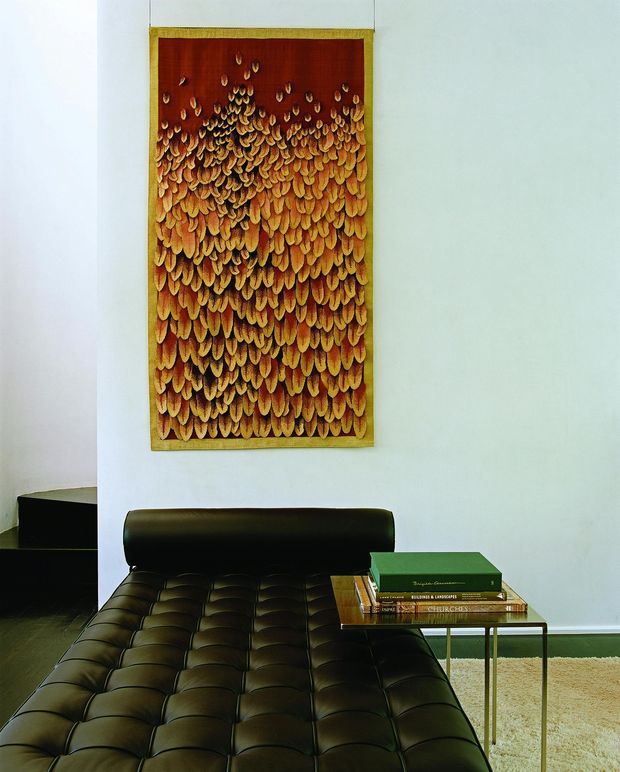 A refined interior vignette featuring a black tufted leather lounge chair or daybed in the foreground, paired with a slim brass-toned side table holding books. A large vertically oriented abstract artwork in warm ochre, amber and deep red tones hangs on a pale wall behind it. The room has dark flooring, minimal furnishings and a restrained, gallery-like composition.