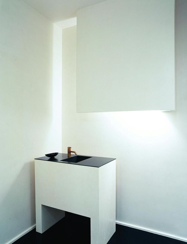 Minimalist bathroom vanity in a compact, bright room. A freestanding white rectangular vanity with an integrated black basin and black stone-like benchtop is paired with a simple bronze tap. A large plain white wall-mounted panel or mirror occupies the upper wall, with concealed lighting beneath it. The room has white painted walls and a very dark floor, creating a stark monochromatic composition.