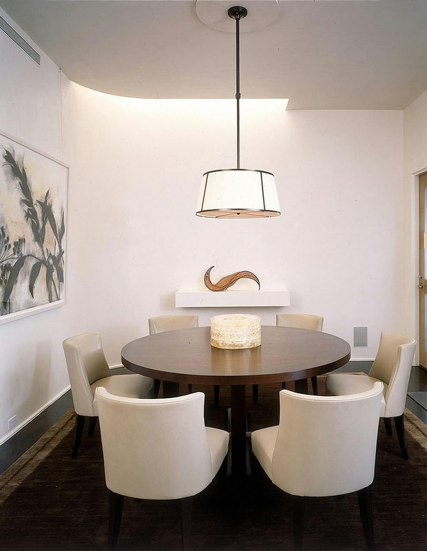 A refined dining room with a large round dark-wood dining table surrounded by upholstered cream dining chairs. The room has pale walls, dark flooring, a suspended drum pendant light, a minimalist floating shelf with sculptural decor, and abstract wall art. A curved ceiling detail and concealed linear air vent add architectural interest.