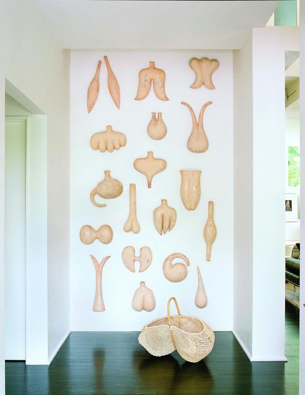 Bright minimalist interior alcove displaying a curated collection of sculptural, pale terracotta or wooden organic forms mounted on a white wall. The niche has dark timber flooring, crisp white walls and ceiling, and a woven basket placed on the floor. Openings at either side reveal portions of adjacent rooms, while soft daylight creates an airy, gallery-like atmosphere.