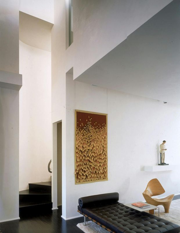 A bright, double-height modern living room with white sculptural walls and a black staircase. The space features a black tufted daybed, a sculptural wooden lounge chair, a pale rug, a small glass coffee table, and a large textured wall artwork. Minimal recessed lighting and clean architectural geometry create an airy, gallery-like atmosphere.