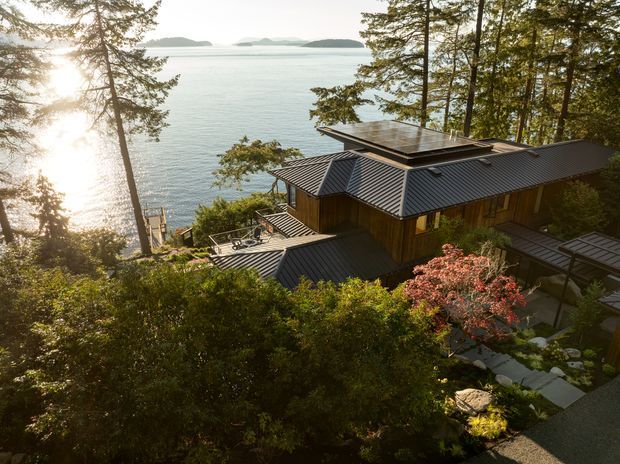 Elevated exterior view of a contemporary timber-clad house nestled among mature trees on a steep, landscaped waterfront site. The building features multiple intersecting dark metal pitched roofs, broad roof overhangs, and a flat rooftop section, with expansive views across calm water toward distant islands. Stone paths, planted garden areas, and outdoor terraces connect the house to the surrounding landscape.