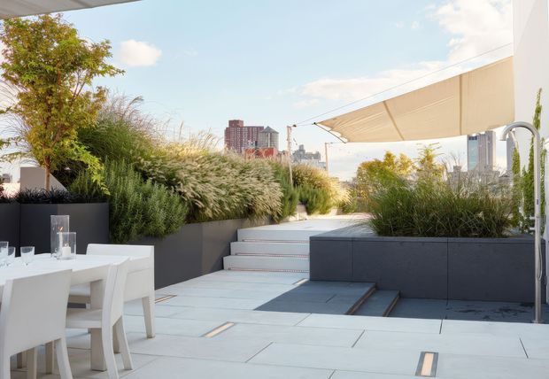 A contemporary rooftop terrace with pale large-format paving, integrated dark raised planters, lush ornamental grasses and trees, and a white outdoor dining setting. Broad retractable shade sails provide shelter, while steps lead between planted levels. Urban high-rise buildings are visible beyond the terrace, creating a landscaped city rooftop atmosphere.